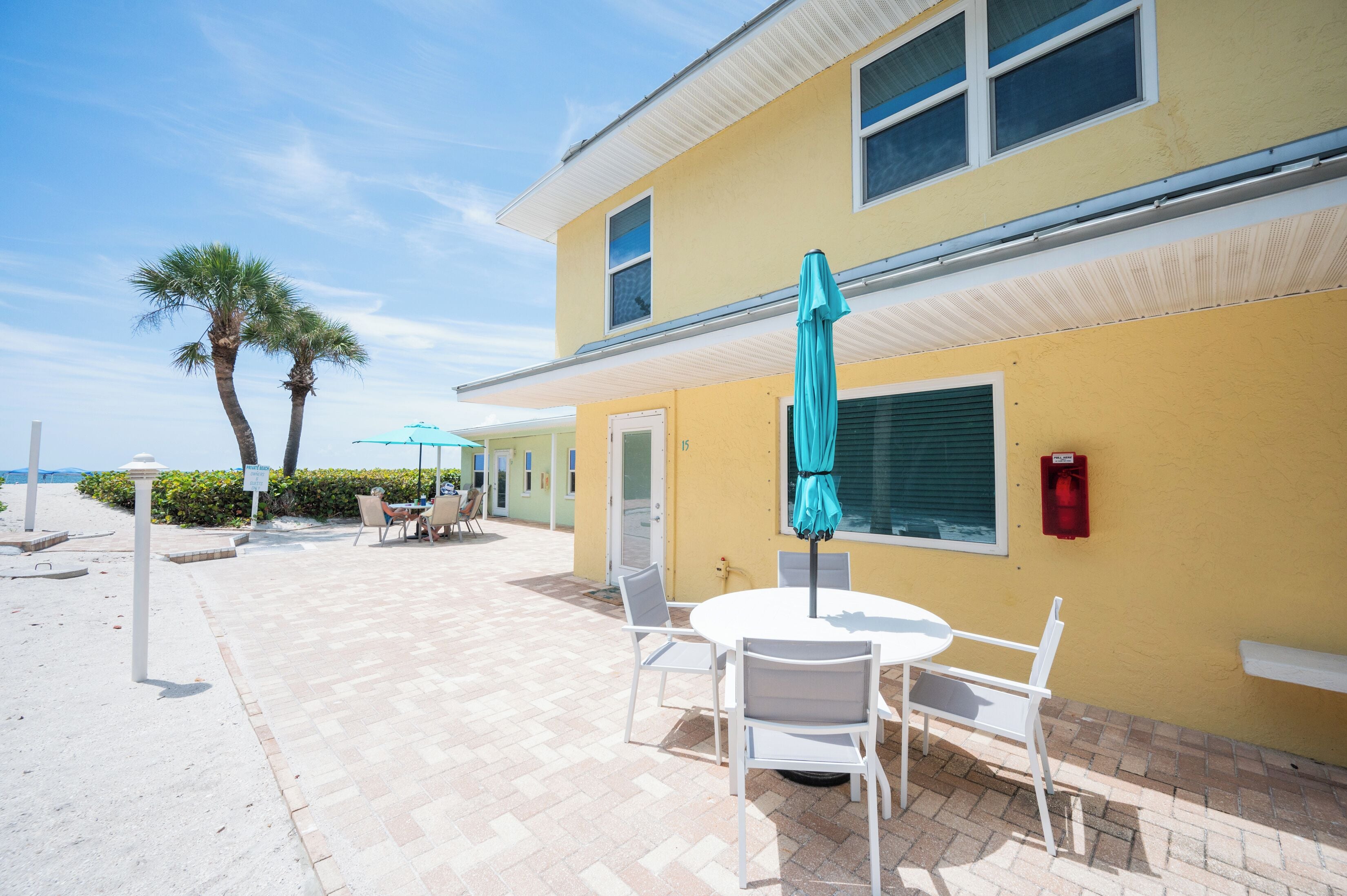 Un edificio amarillo con un patio que tiene una mesa redonda y sillas, una sombrilla azul y vista a la playa.