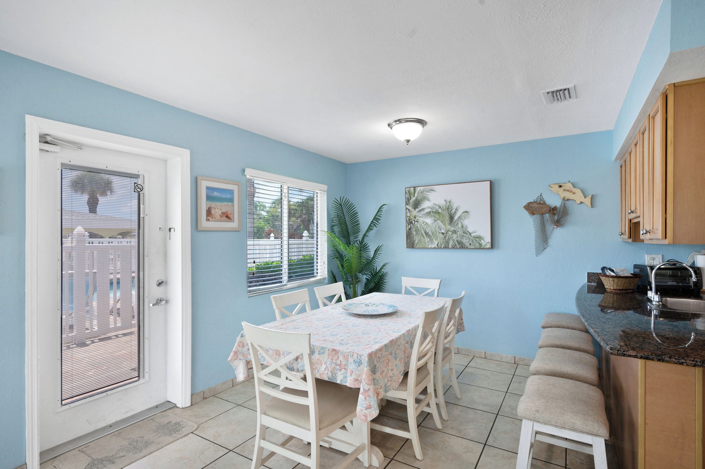 A dining area with a table and chairs, a window with blinds, a potted plant, and a kitchen counter with a sink.