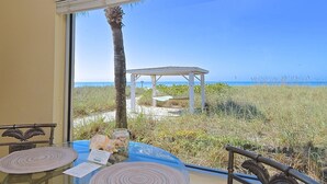 Una habitación con un ventanal grande con vistas a la playa, una mesita con un mantel individual y una silla con respaldo decorativo.