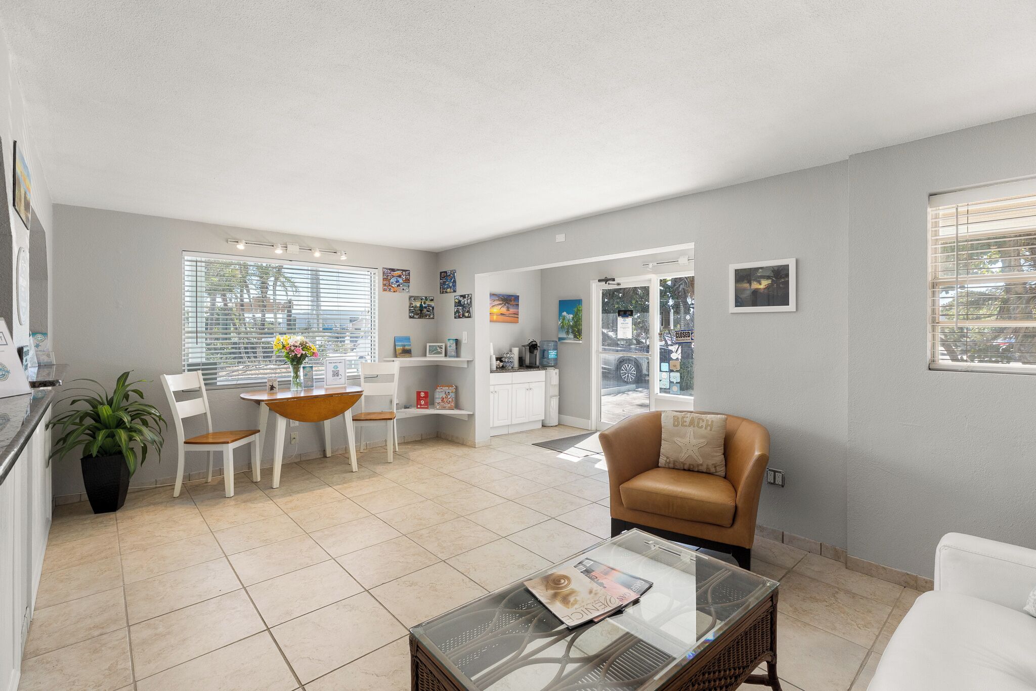 Un salón moderno con una mesa de centro de vidrio, muebles blancos, un sofá de cuero marrón y una cocina visible a través de una puerta abierta.