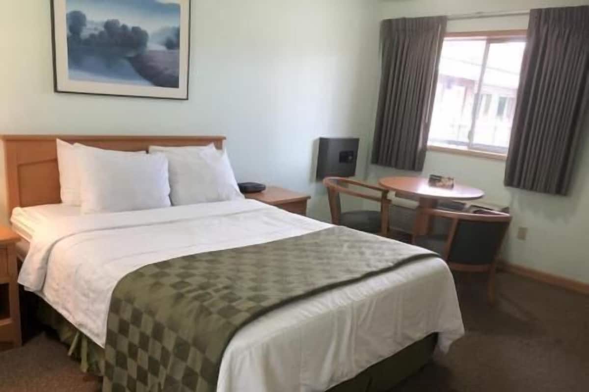 Ett hotellrum med en säng, ett skrivbord, en stol, ett fönster med gardiner och en tavla på väggen.