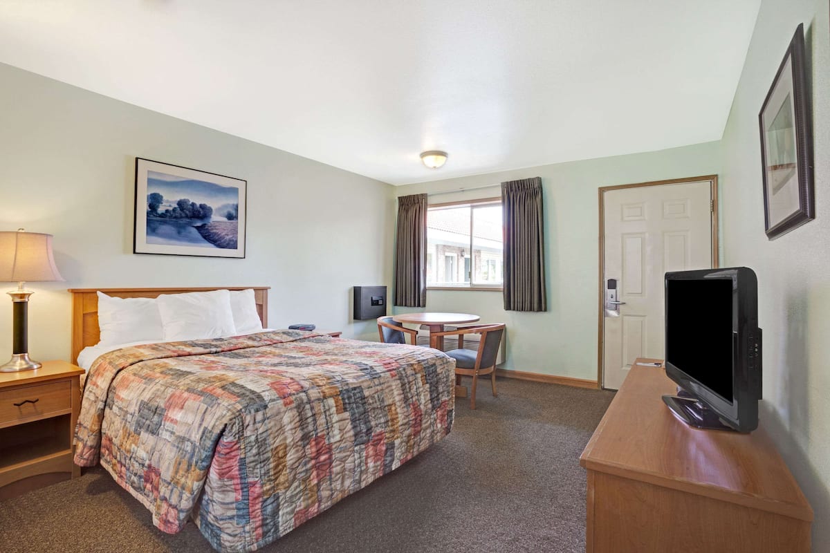 Habitación de hotel con una cama, un escritorio con televisor, una silla, una lámpara, un cuadro en la pared y una ventana con cortinas.