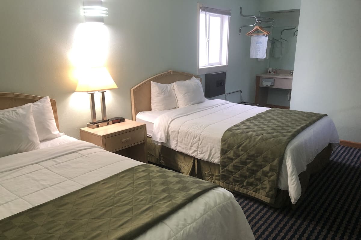 Habitación de hotel con dos camas, una mesita de noche, una lámpara, una ventana, un espejo y un baño con toallero.