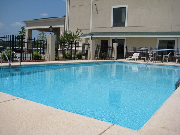 Una piscina rectangular con agua azul cristalina, rodeada por una terraza de baldosas color beige. Se observan sillas de descanso y una valla de metal negro alrededor de la piscina.