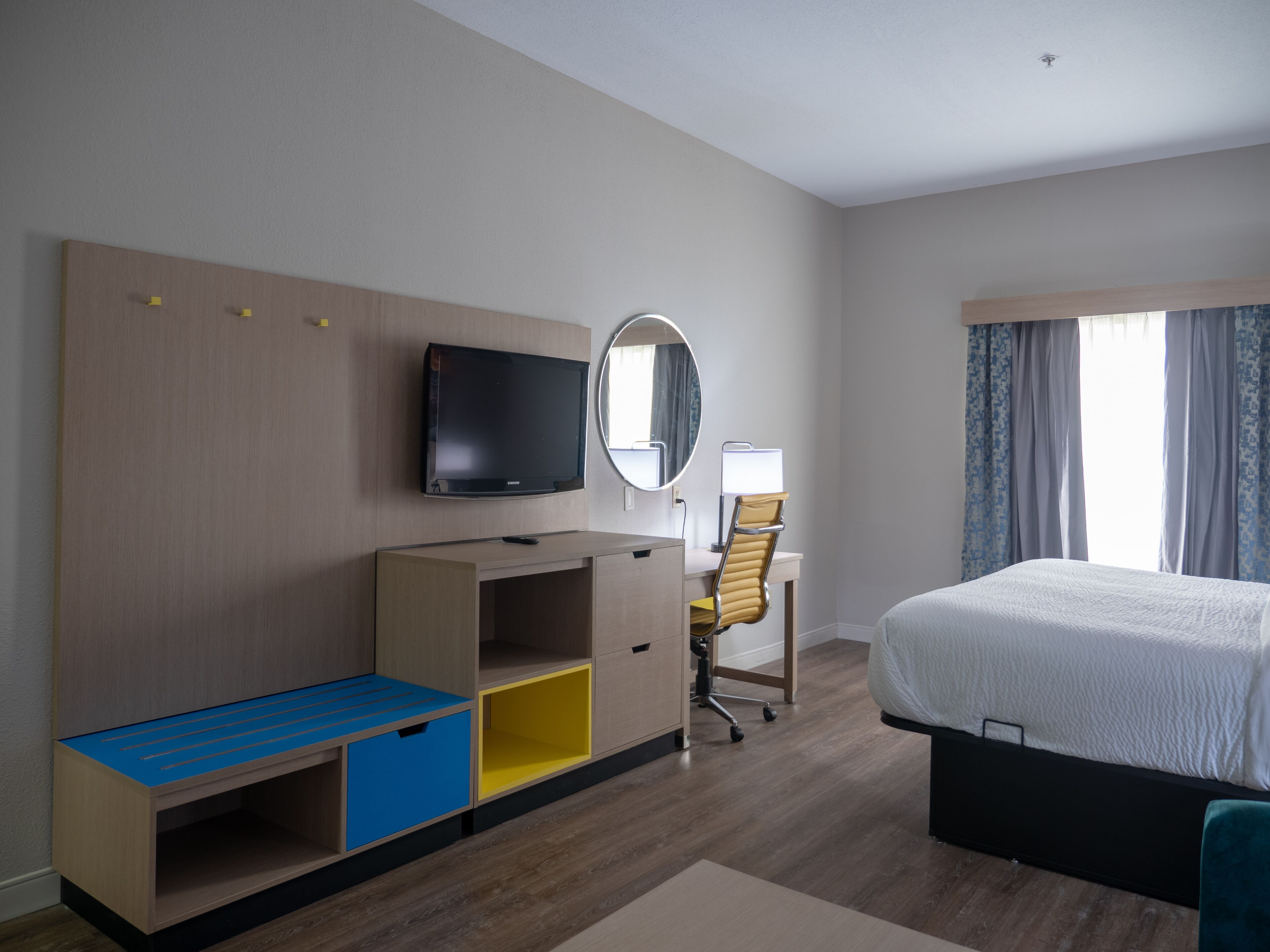 Habitación de hotel con cama, escritorio, silla, televisor y espejo.