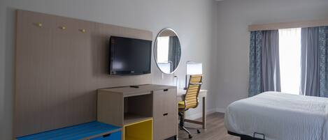 Habitación de hotel con cama, escritorio, silla, televisor y espejo.