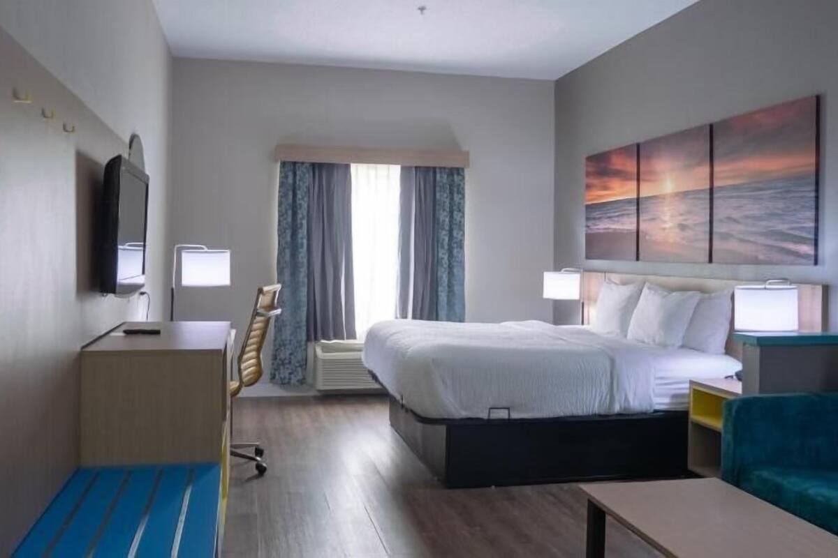 Ein Hotelzimmer mit einem Bett, einem Schreibtisch mit Stuhl, einem Fernseher, einem Fenster mit Vorhängen, einem Bild und einem blauen Sofa.