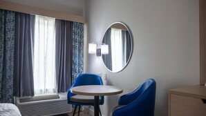 Una habitación de hotel con cama, una mesa redonda, dos sillas azules, un calefactor, un espejo y una ventana con cortinas.