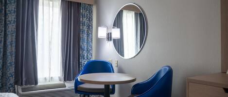 Una habitación de hotel con cama, una mesa redonda, dos sillas azules, un calefactor, un espejo y una ventana con cortinas.