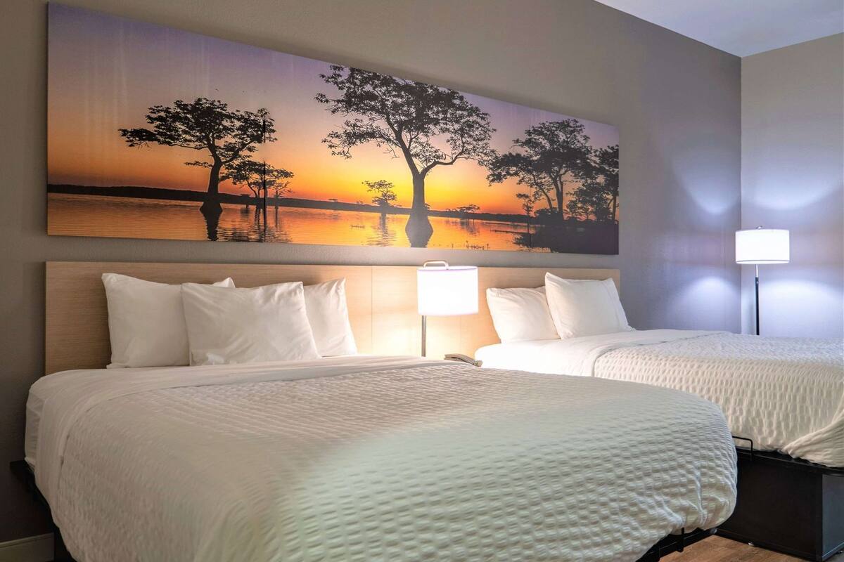 Une chambre d’hôtel avec deux lits, un grand tableau représentant un coucher de soleil et une table de chevet avec une lampe.