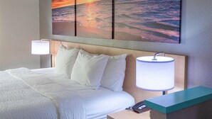 Habitación de hotel con una cama, ropa de cama blanca, una mesita de noche con una lámpara y una obra de arte colgada en la pared que representa un atardecer sobre el océano.