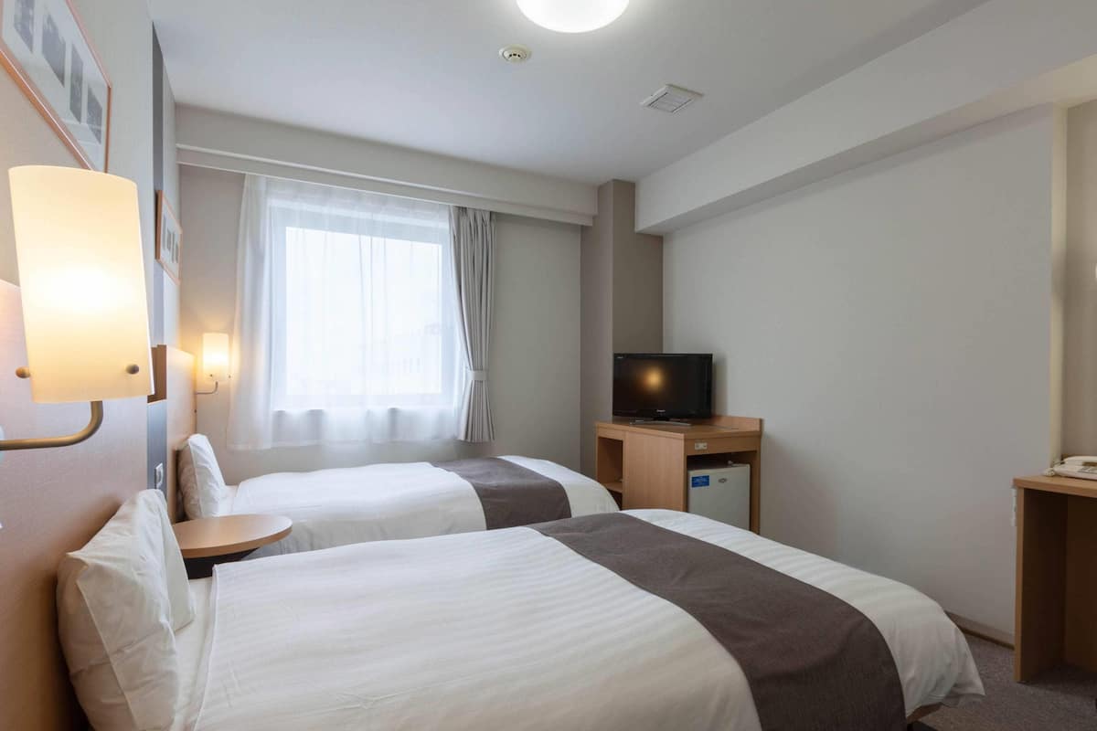A hotel room with two beds, a television on a wooden stand, a desk with a phone, and a window with curtains.