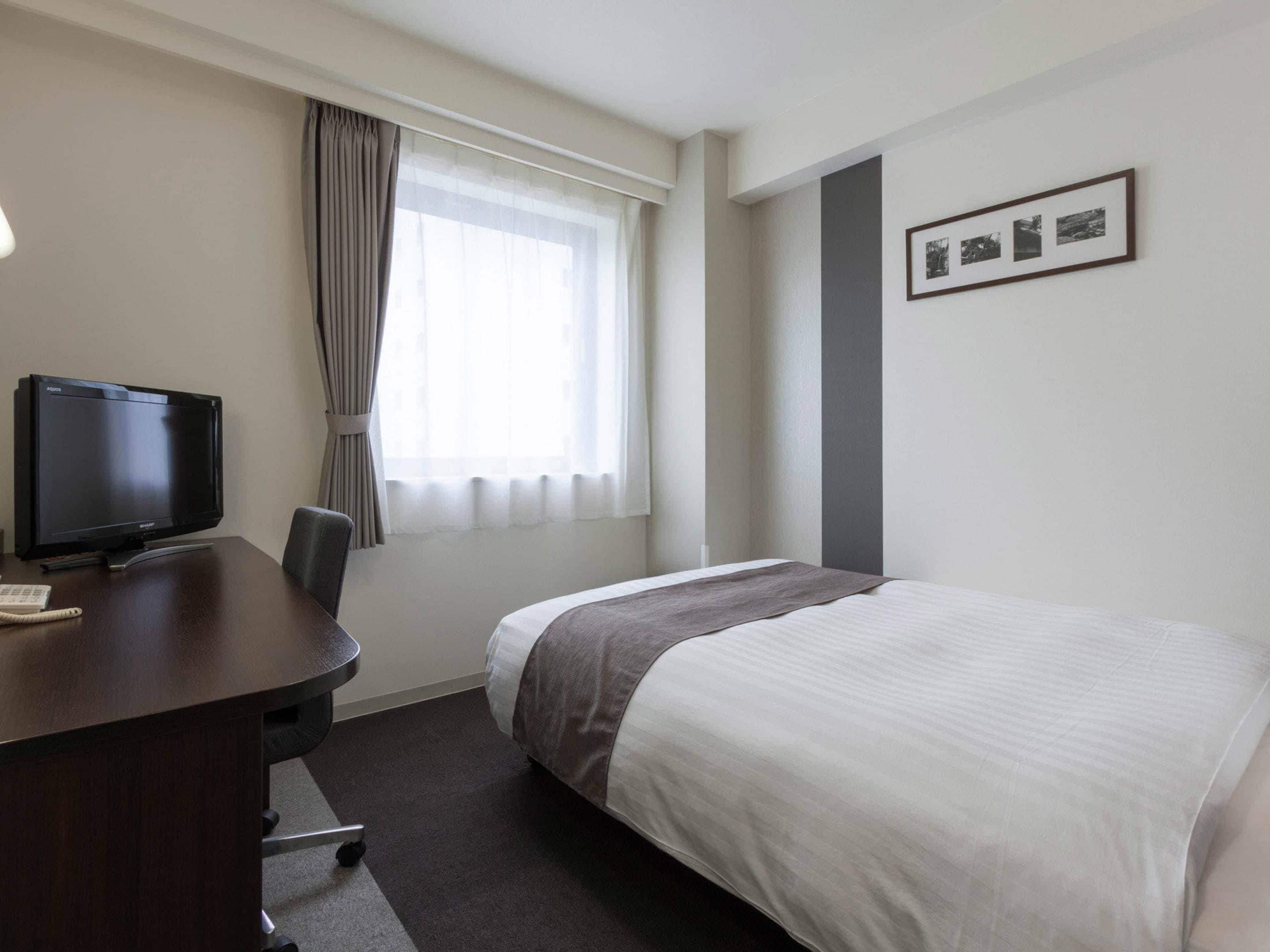 A hotel room with a bed, a desk with a television, a chair, a window with curtains, and a framed picture on the wall.