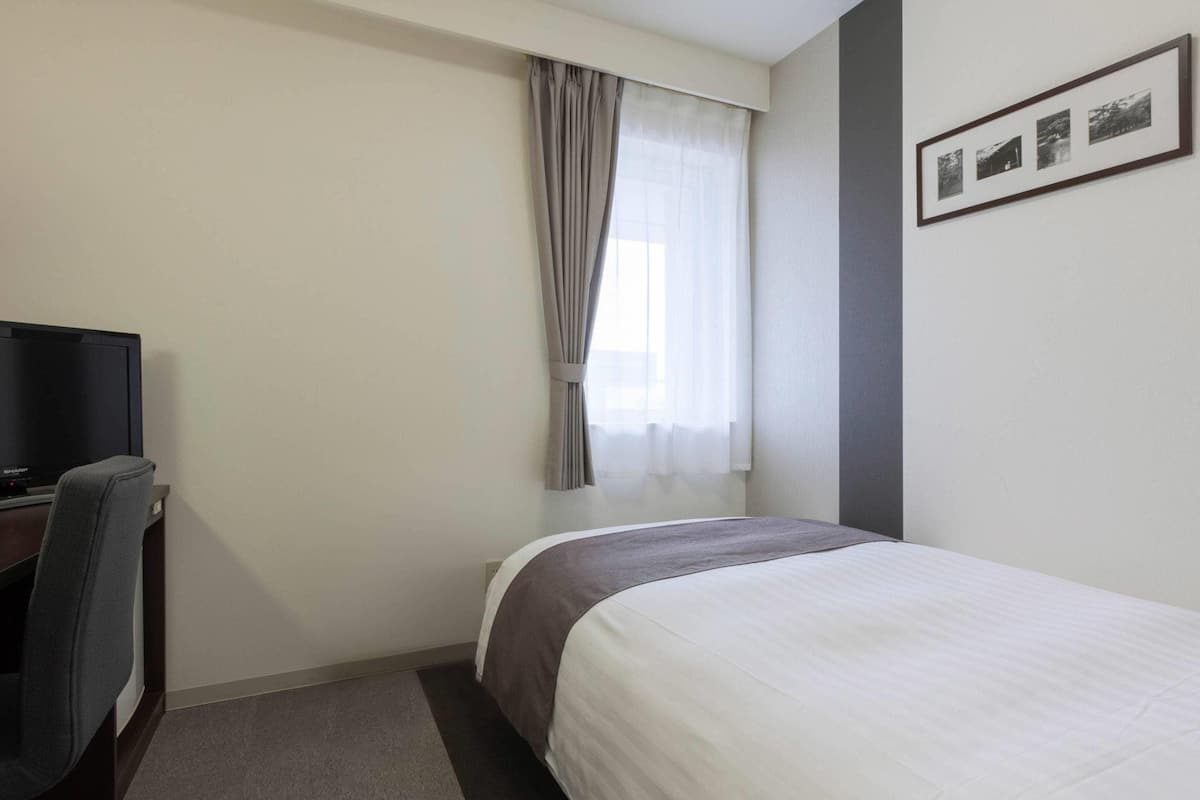 A hotel room with a bed, a desk with a computer, a chair, a window with curtains, and a framed photo on the wall.