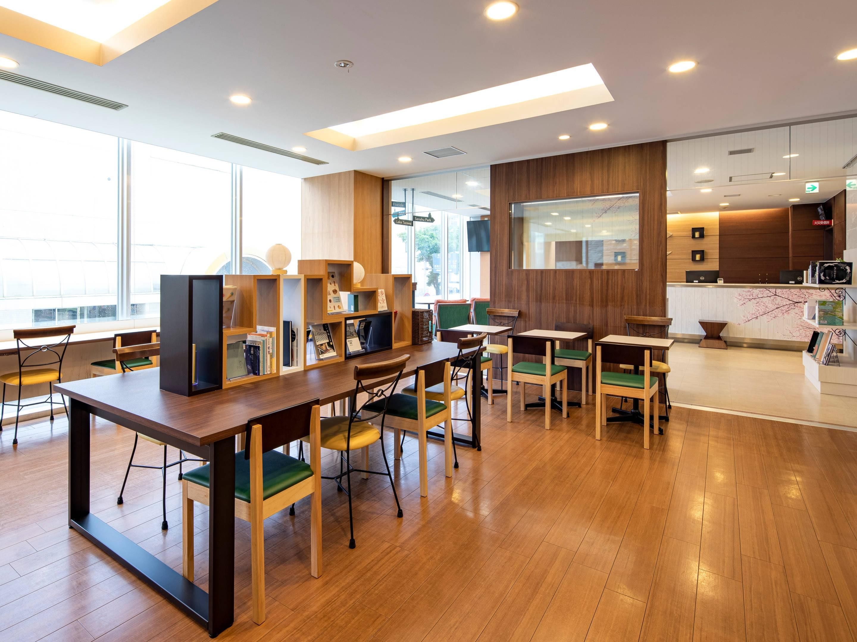 A modern library with wooden tables and chairs, bookshelves, and a large window providing natural light.