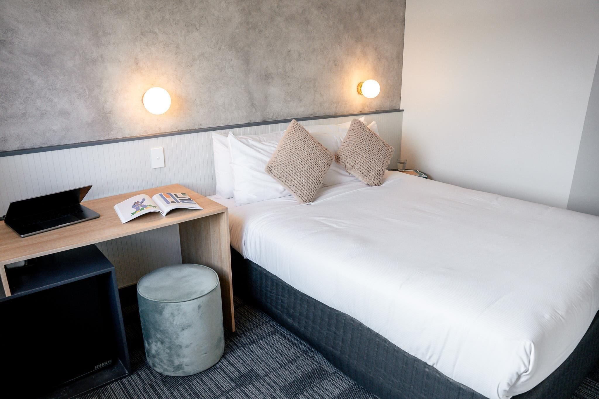 Habitación de hotel con una cama, un escritorio con una computadora portátil, un libro y un cesto de basura.