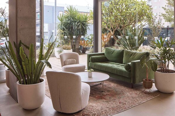 A modern living room with a green sofa, a white armchair, and a coffee table. There are several potted plants and a large window providing a view of the city. - Hotel TOTTO Wollongong (Wollongong)