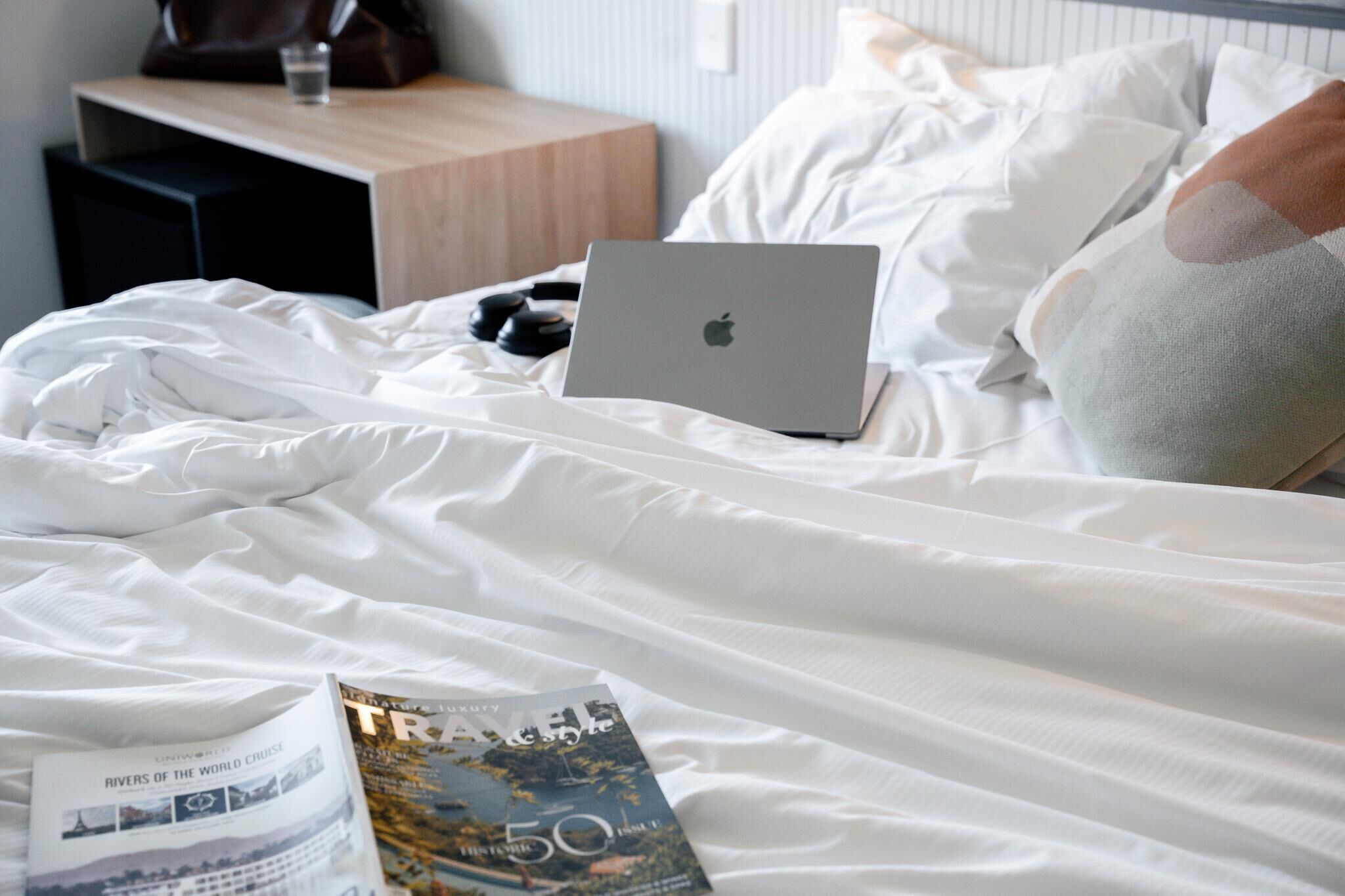 Una cama bien tendida con una computadora portátil, una revista y un vaso de agua sobre una mesita de noche de madera.
