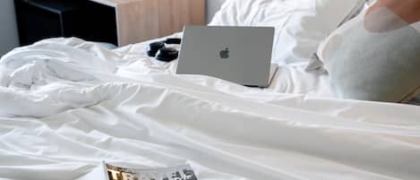 Una cama bien tendida con una computadora portátil, una revista y un vaso de agua sobre una mesita de noche de madera.