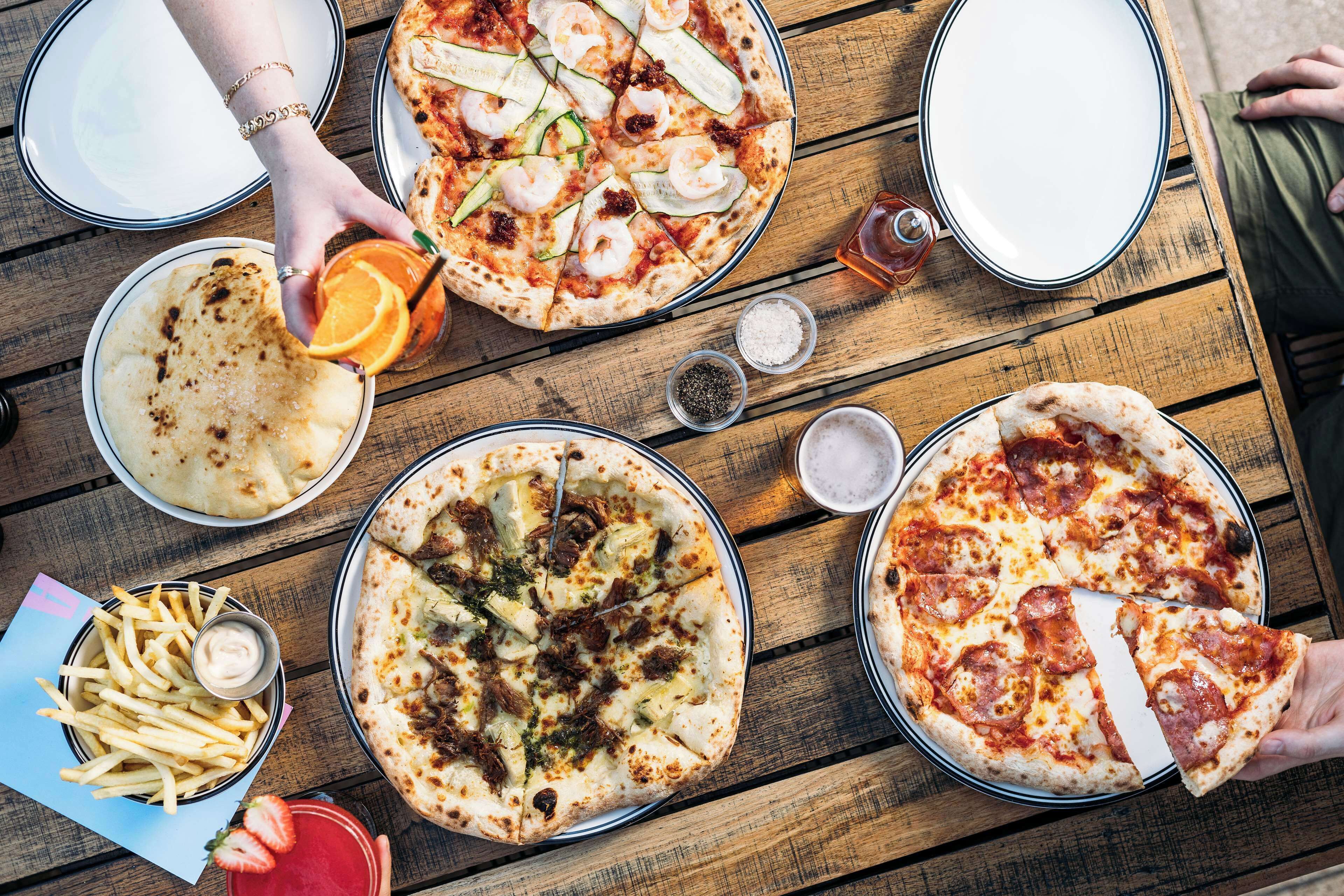 A wooden table with various pizzas, a bowl of fries, and a glass of orange juice.