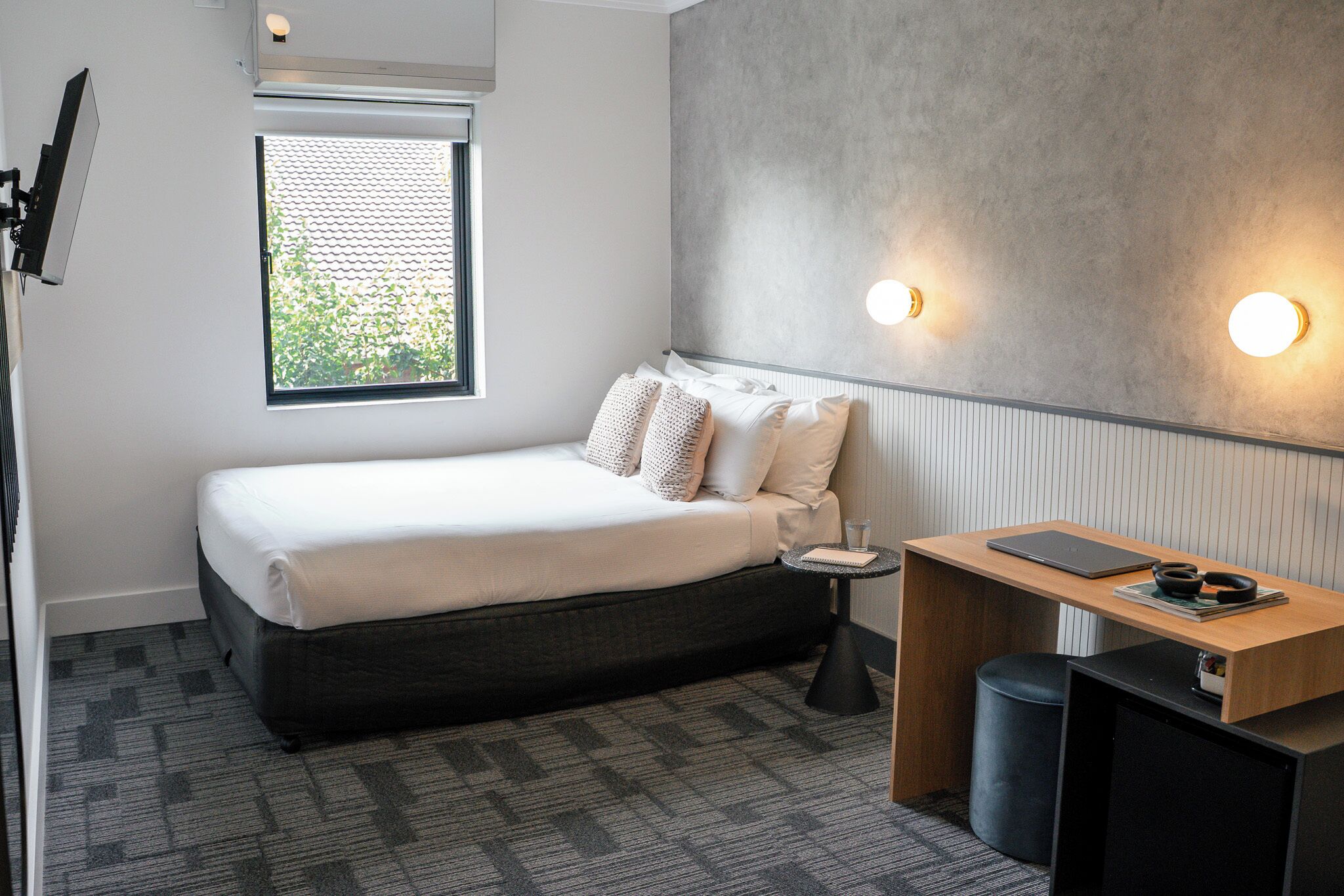 A modern hotel room with a bed, a small desk, a chair, a lamp, and a window with blinds.