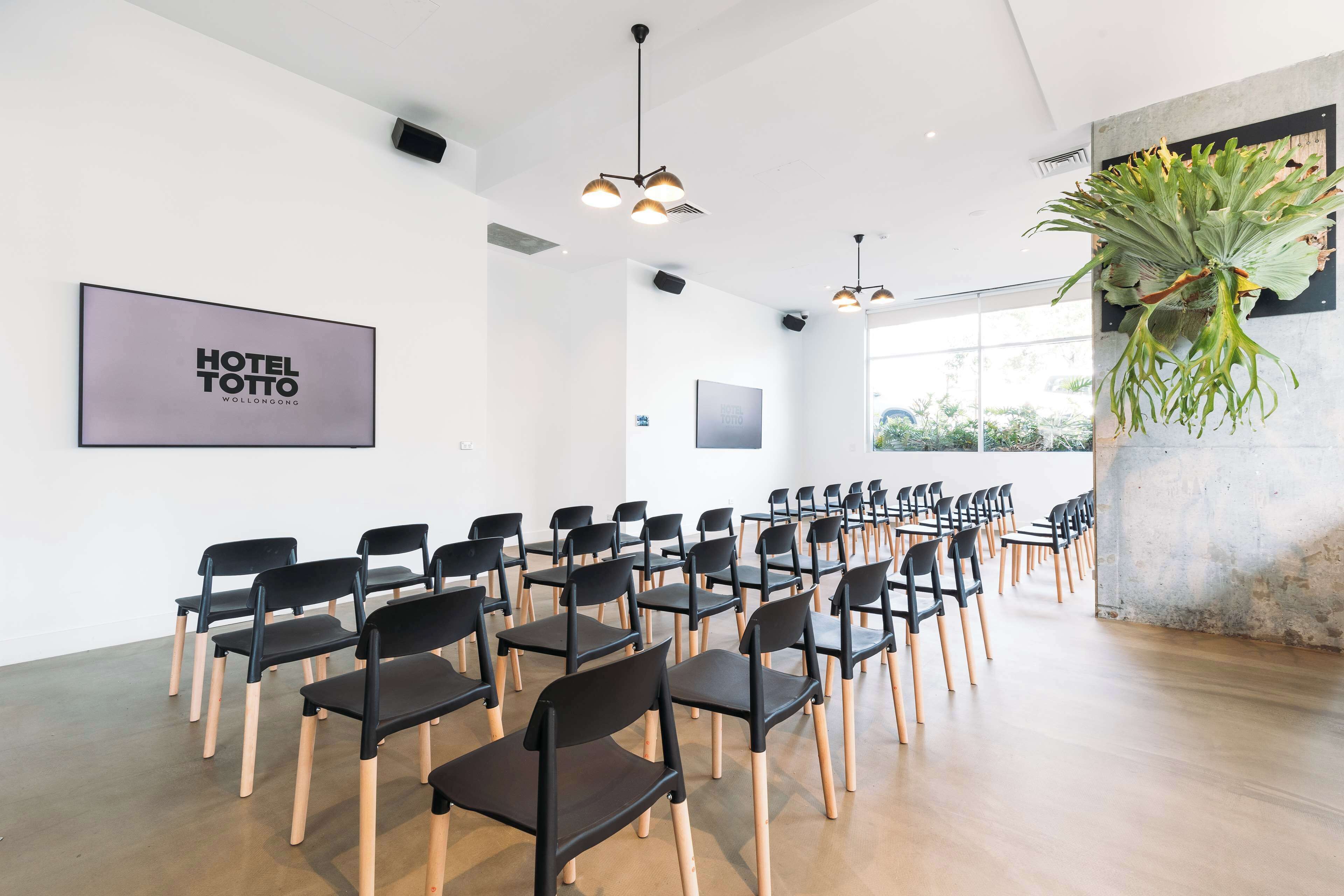 Een congreszaal met rijen zwarte stoelen en houten vloeren, een grote plant en een 'HOTEL TOTTO' bord aan de muur.