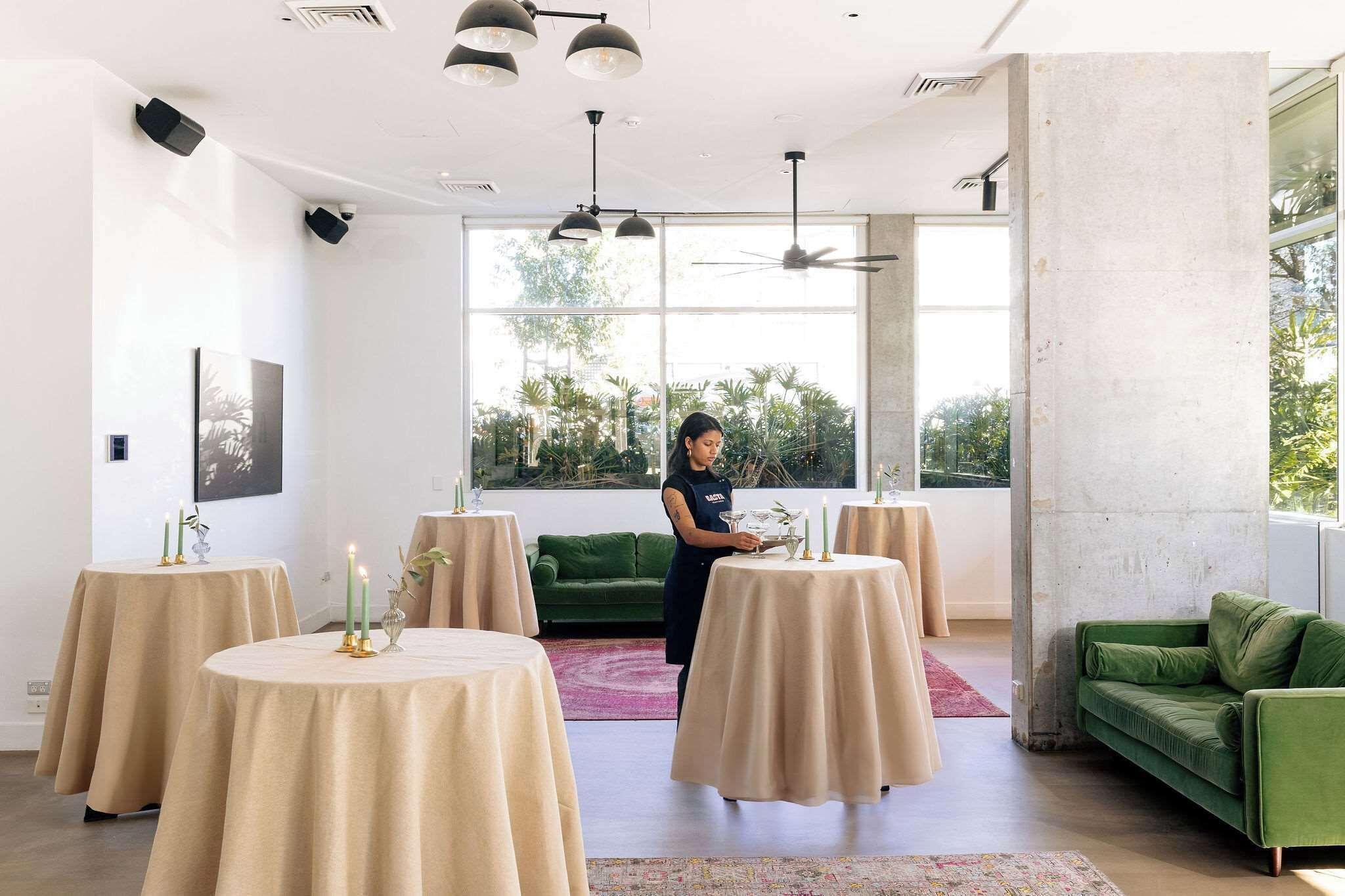 Un interior moderno con mesas redondas cubiertas con tela color beige, un sofá verde y una persona ordenando objetos en una mesa.