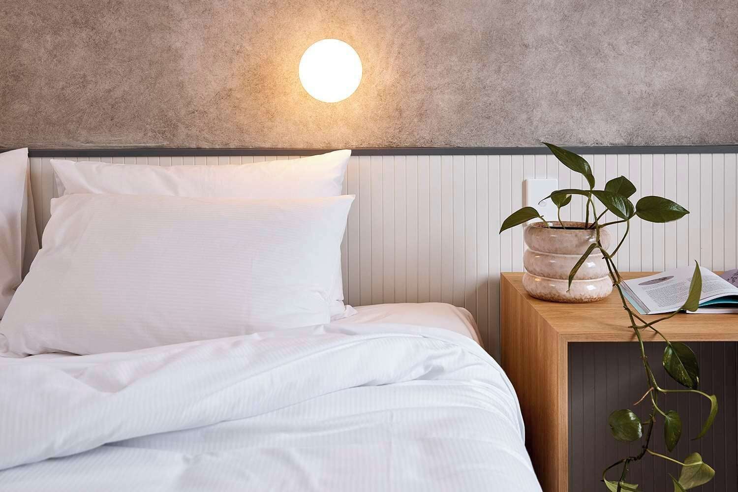 A neatly made bed with white linens and pillows, a round wall-mounted light, a wooden nightstand with a potted plant and an open book, and a textured wall behind the bed.
