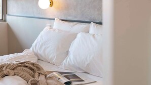 Un lit bien fait, avec des draps blancs et une couverture tricotée de couleur beige, un magazine posé sur le lit et un luminaire fixé au mur.