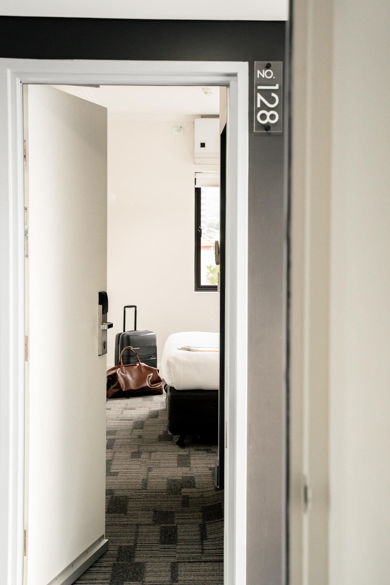 A hotel room with a bed, a suitcase, a handbag, and a wall-mounted air conditioning unit.