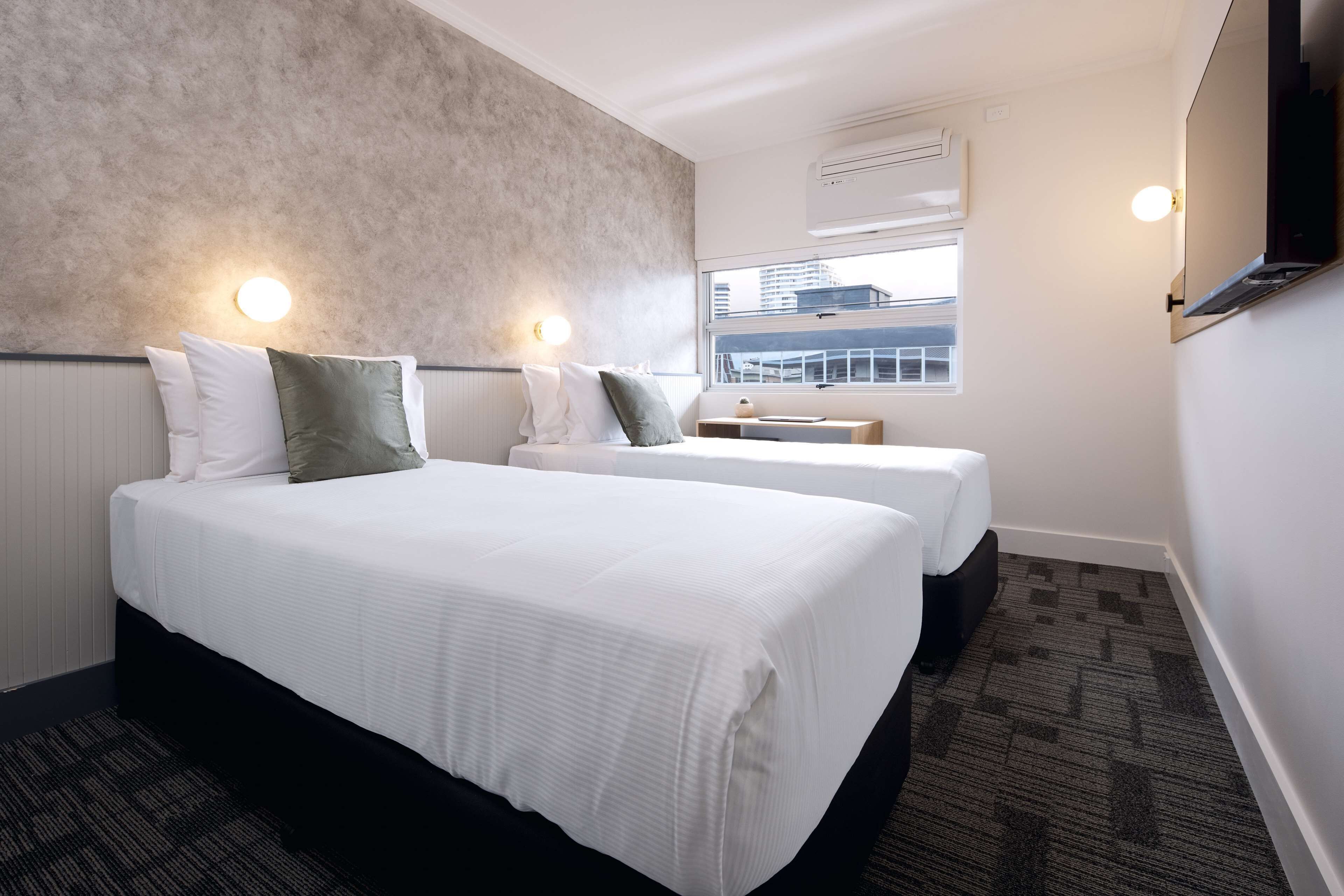 Ein Hotelzimmer mit einem großen Bett, zwei Nachttischen, einem Fenster mit Blick auf Gebäude und einer Wand-Klimaanlage.