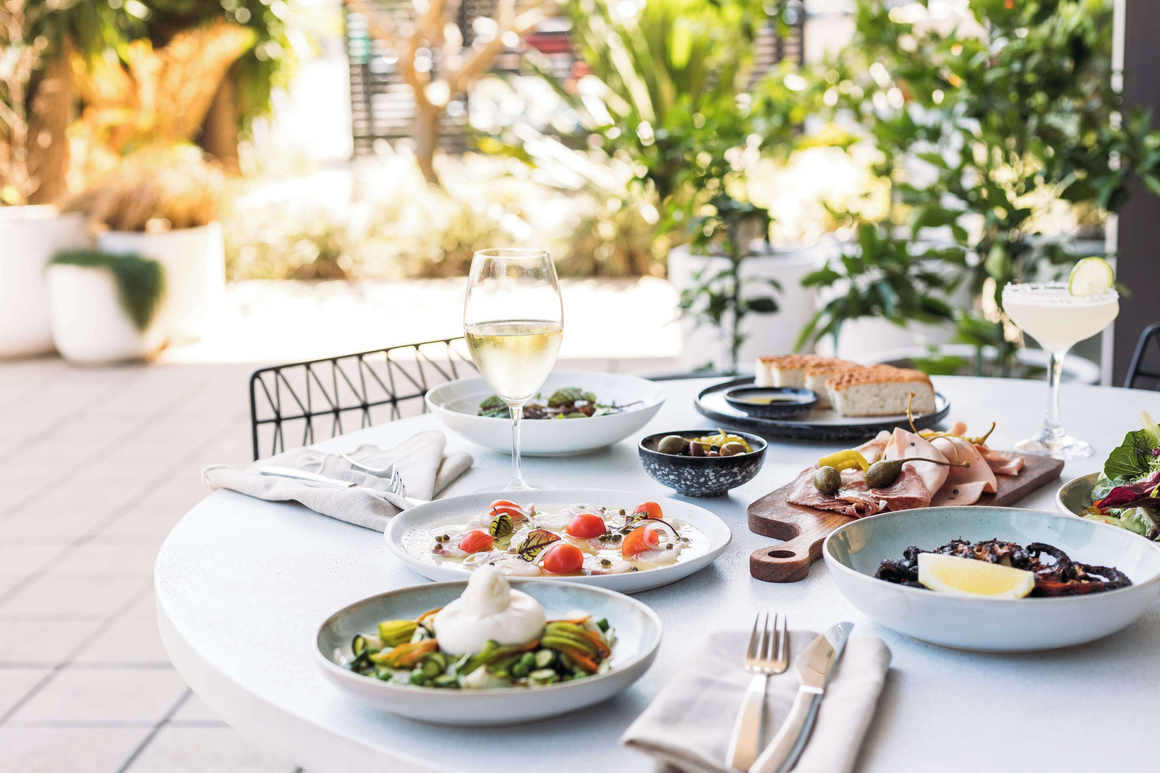 A table set with various dishes, including a salad, a bowl of soup, and a plate of meat, in an outdoor dining area with plants in the background.