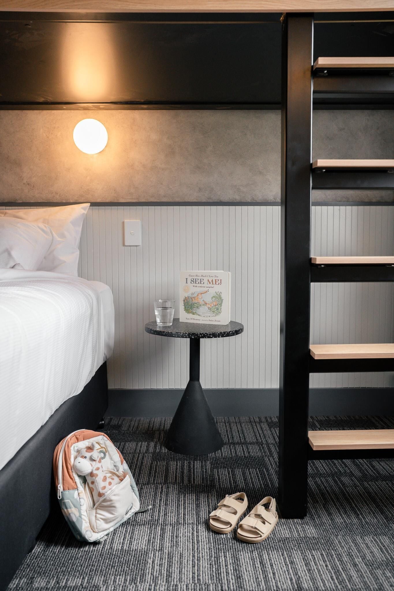 A hotel room with a bed, a small table with a book and a glass, a backpack, and a pair of slippers.