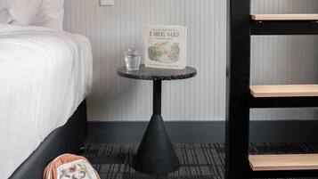 A hotel room with a bed, a small table with a book and a glass, a backpack, and a pair of slippers.