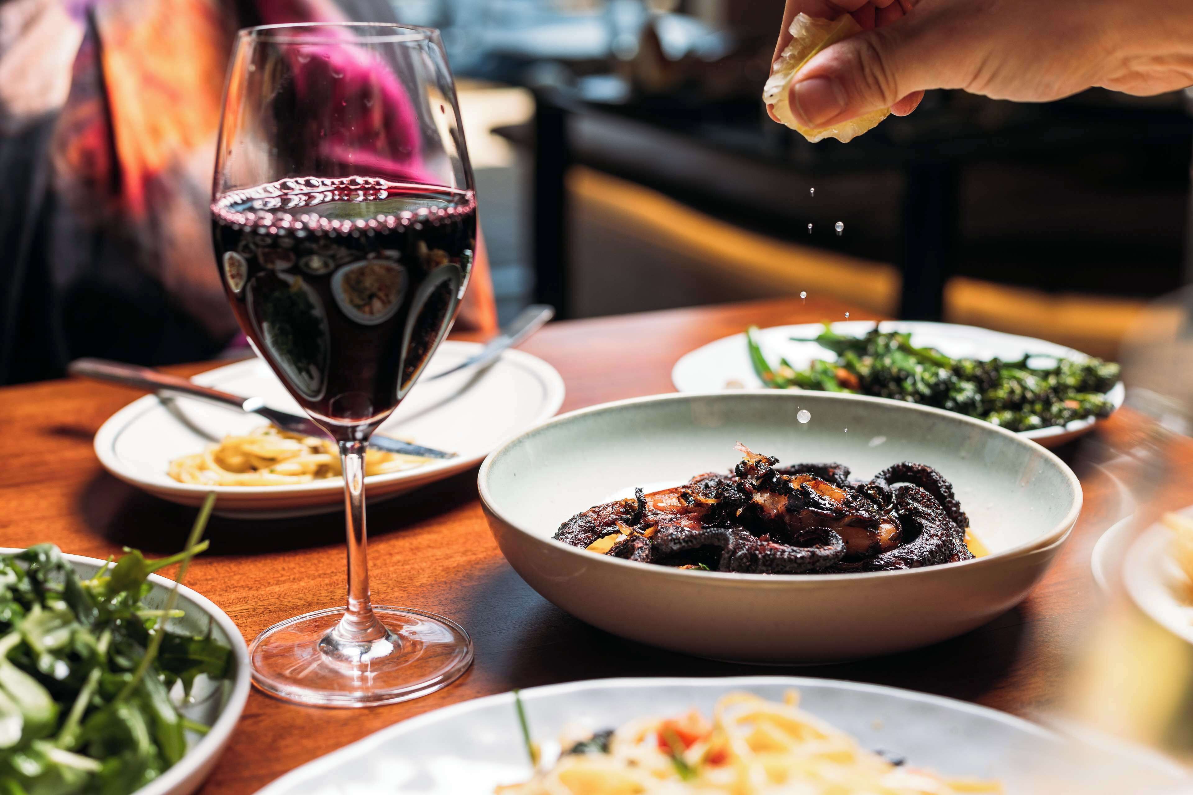 A dining table with various dishes, a glass of red wine, and a hand sprinkling seasoning.
