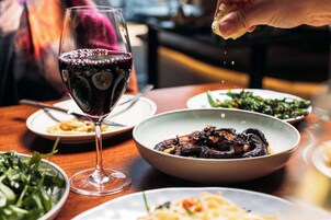 Uma mesa de jantar com vários pratos, um copo de vinho tinto e uma mão polvilhando tempero.