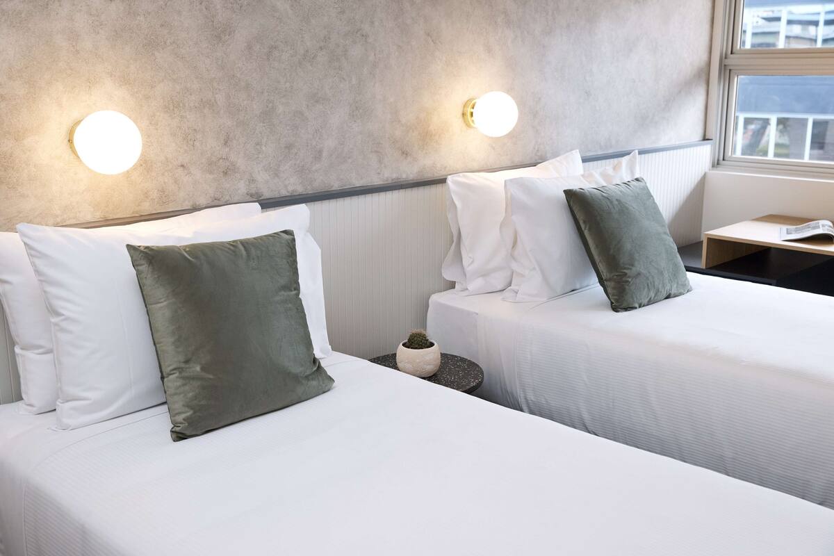 Ein Hotelzimmer mit zwei Betten, weißer Bettwäsche, grünen Kissen, einem kleinen Topfpflanzen und einem Nachttisch mit einem Buch.