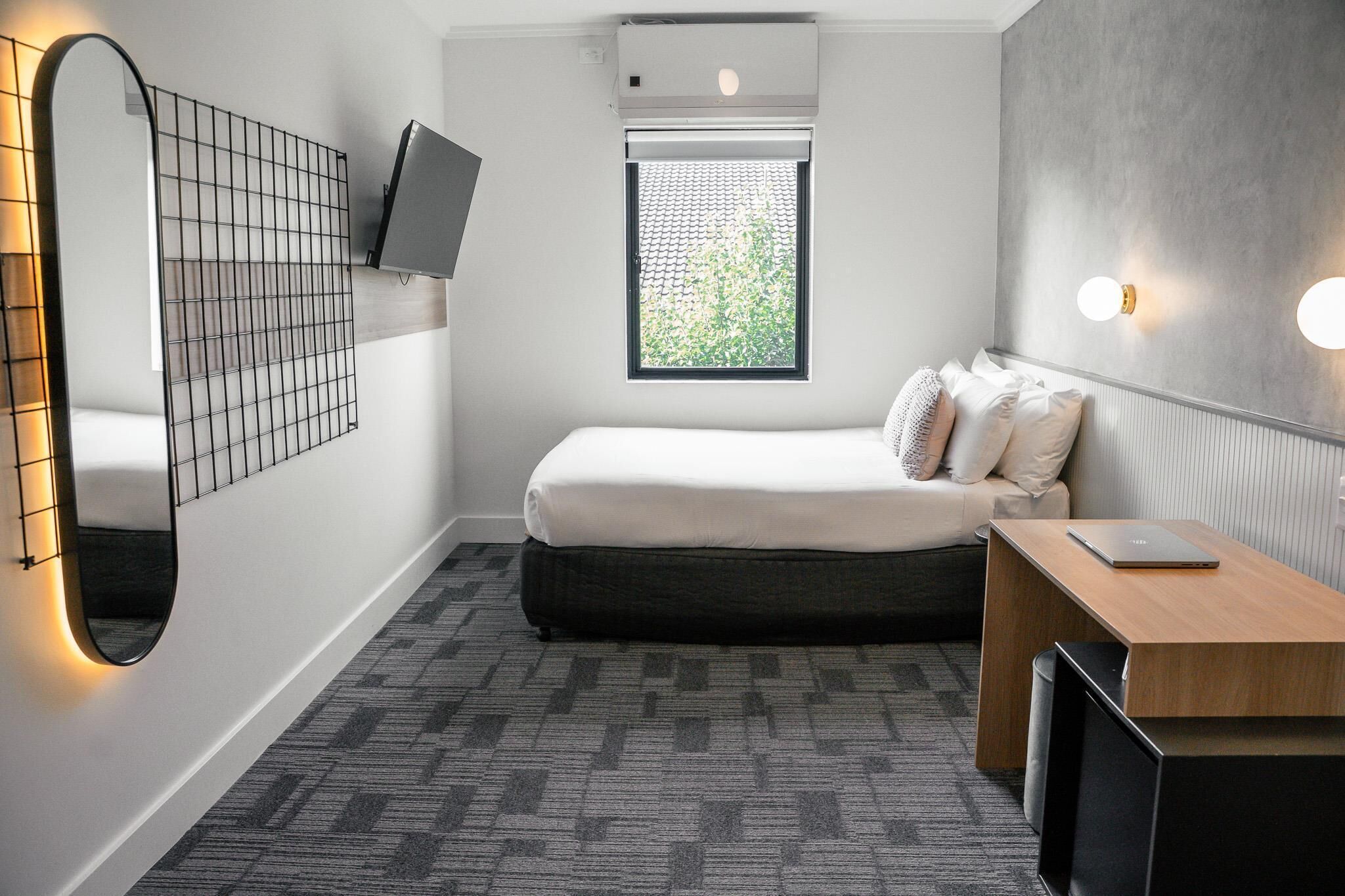 A compact hotel room with a bed, a desk, a television, a window with a view of greenery, and a wall-mounted mirror.