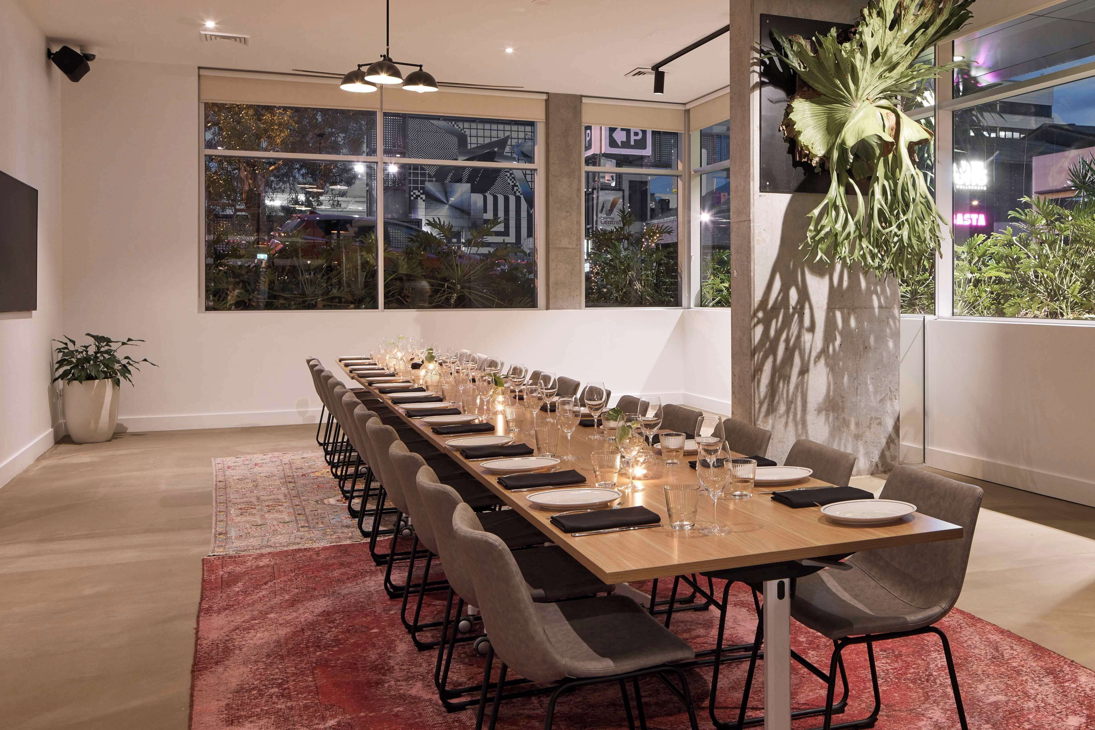 A long conference table set for a meeting with chairs, a potted plant, and a city view through large windows.