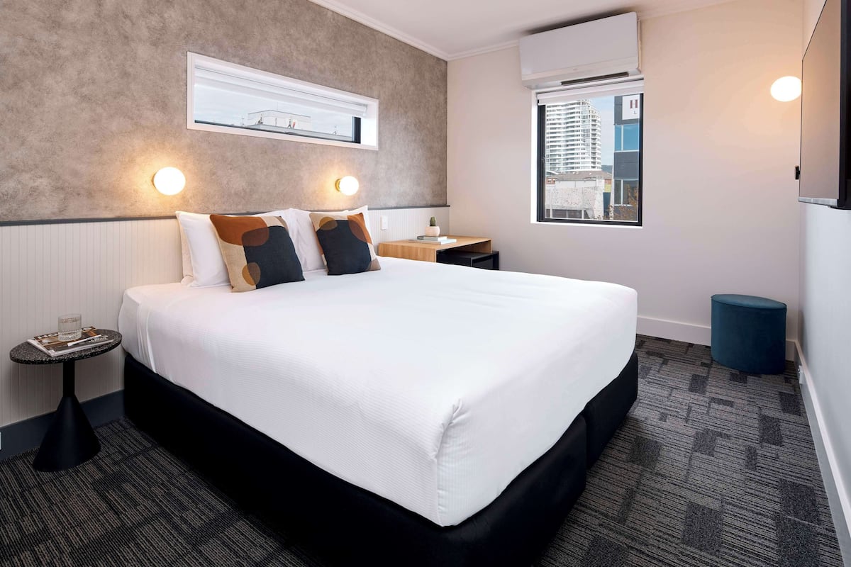 Ein modernes Hotelzimmer mit einem großen Bett, einem kleinen Beistelltisch, einem Nachttisch, einem Fenster mit Blick auf Gebäude und einer wandmontierten Klimaanlage.