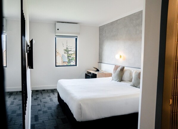 A hotel room with a large bed, a desk with a lamp, a window with a view of buildings, and an air conditioning unit. - Hotel TOTTO Wollongong (Wollongong)