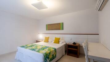 Quarto de hotel com cama, escrivaninha, cadeira, mesa de cabeceira e uma obra de arte na parede.