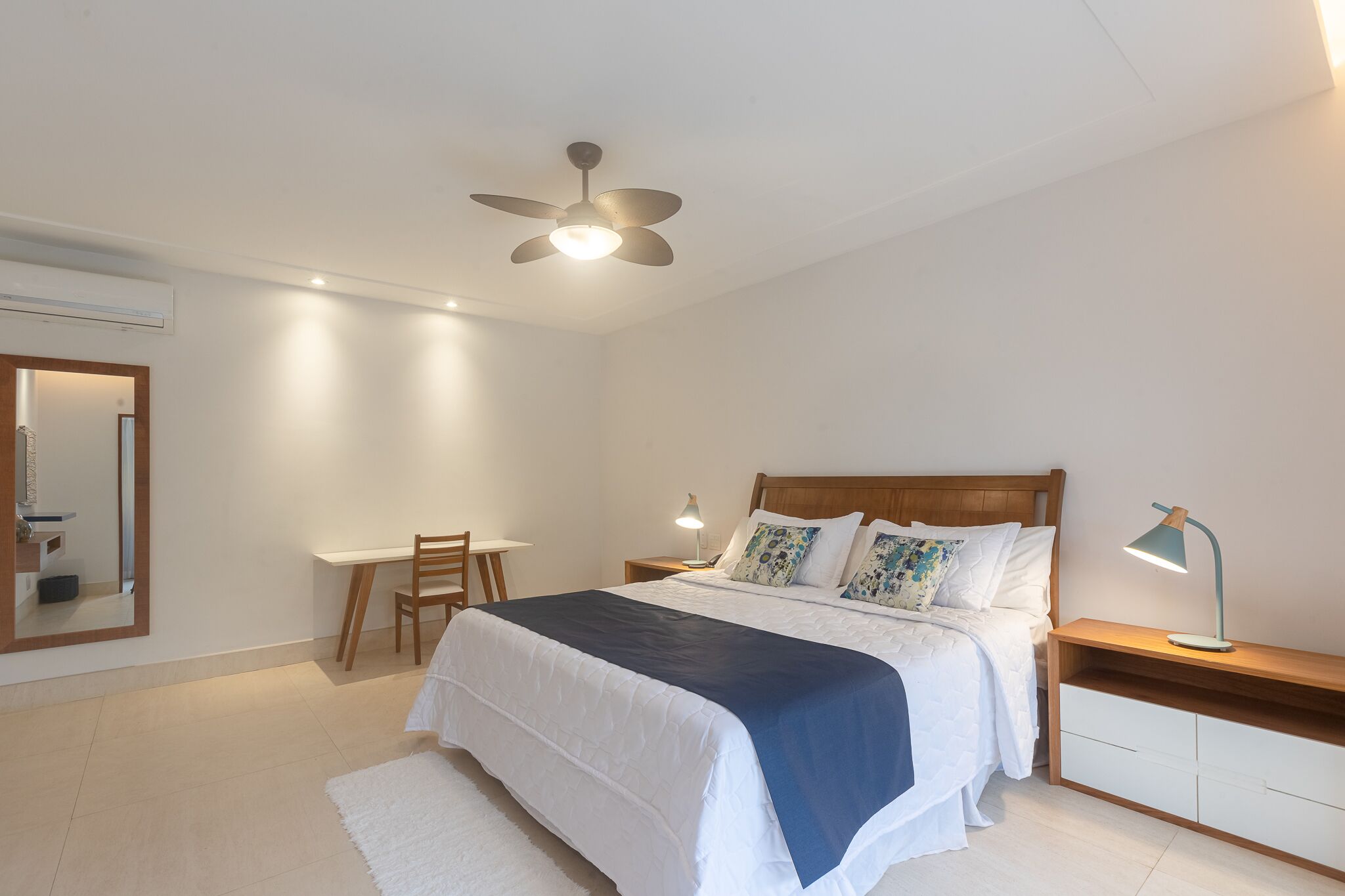 A bedroom with a bed, bedside tables, a desk, a chair, a ceiling fan, and a mirror.