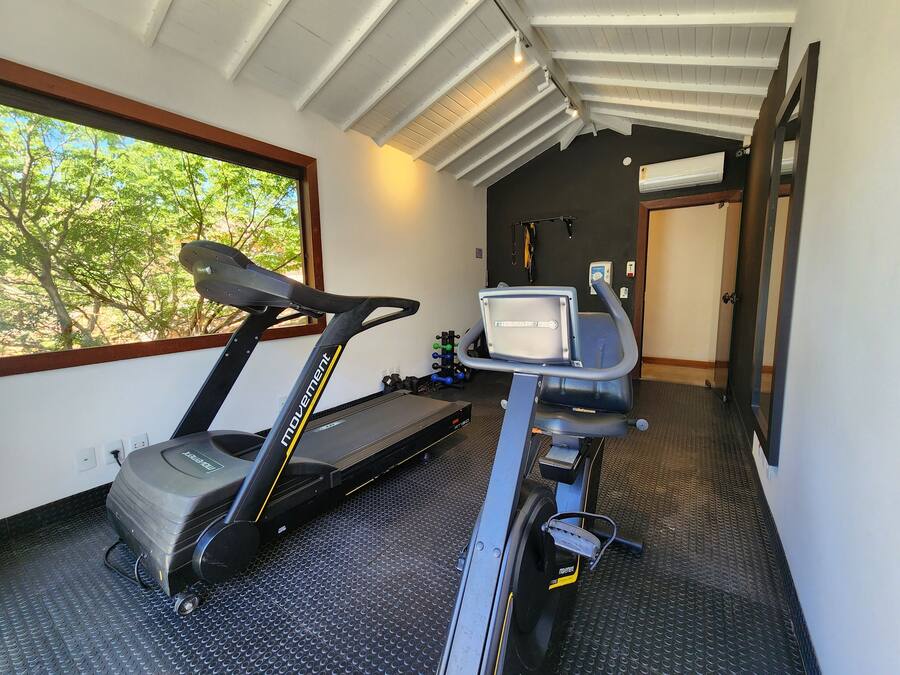 Un gimnasio en casa con una cinta de correr y una máquina elíptica, una ventana con vistas a árboles, y una puerta que da a otra habitación.
