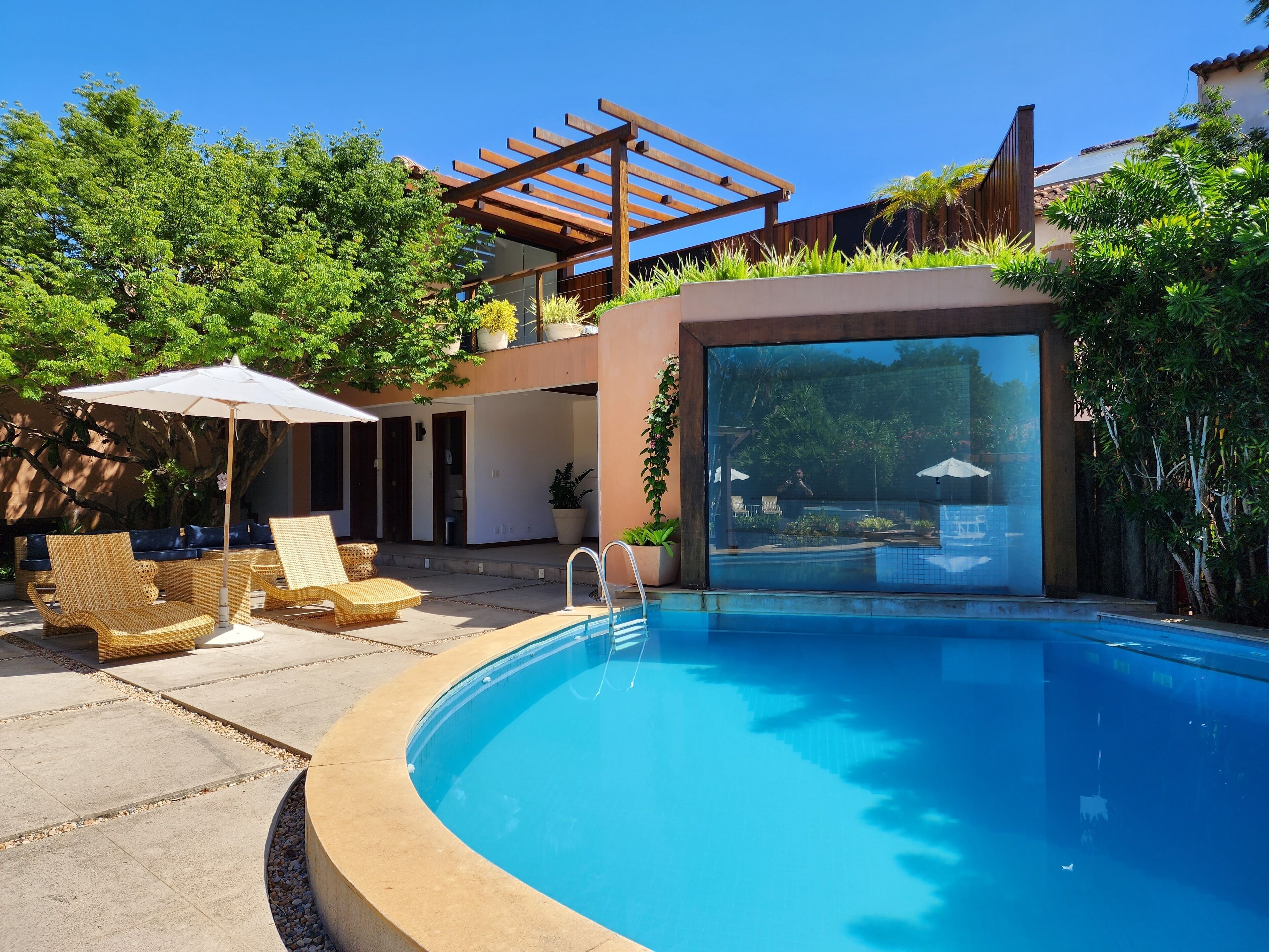 Un área de piscina moderna al aire libre con una pérgola, sillones de descanso y vista a la piscina a través de un muro de cristal.