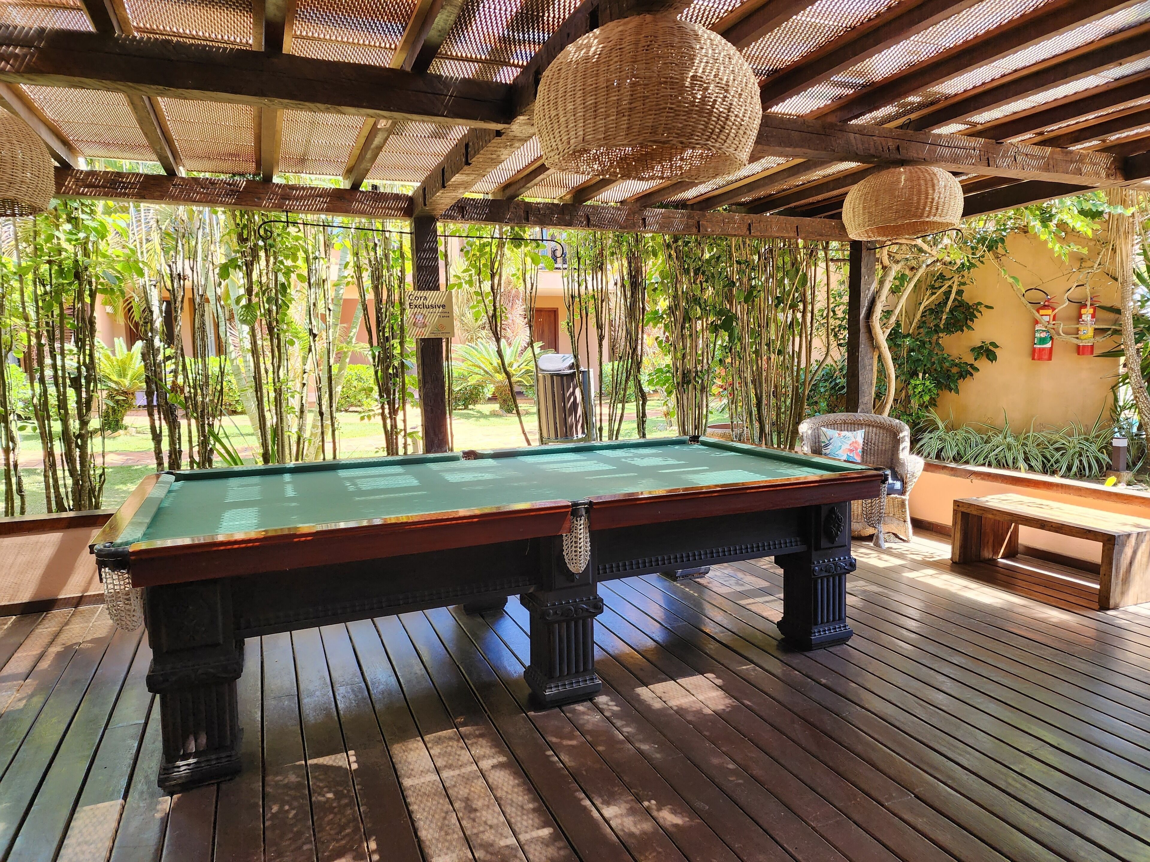 A pool table in a covered outdoor area with a wooden deck, surrounded by bamboo plants and a wicker ceiling.