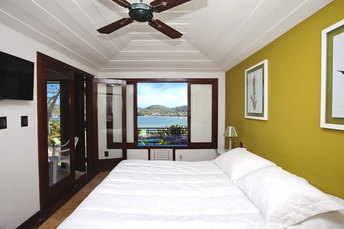 A bedroom with a large bed, a ceiling fan, a window with a view of a body of water and hills, and a television on the wall.