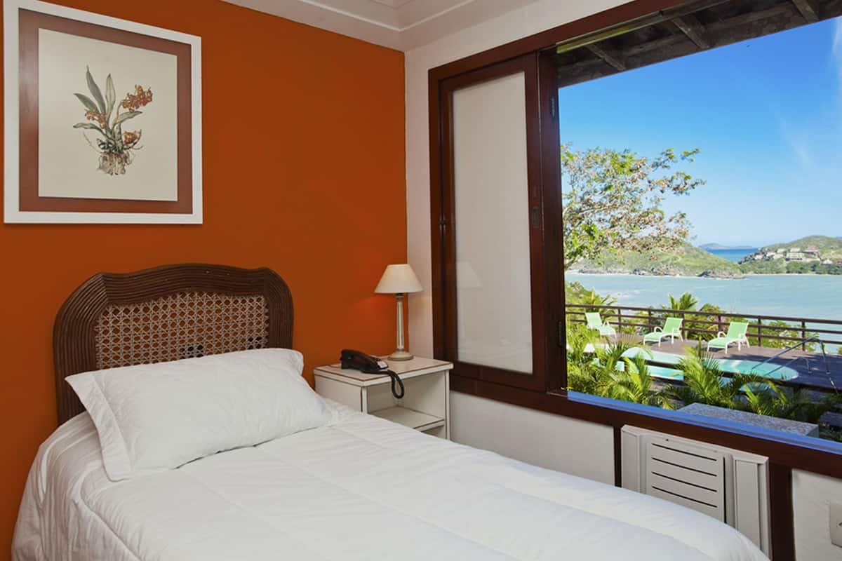 A hotel room with a bed, a nightstand with a lamp, a framed picture on the wall, and a view of the ocean and hills.