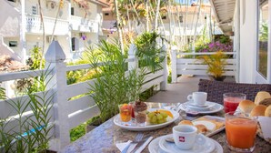A breakfast table with a variety of food items, including fruit, bread, and a glass of juice, set on a balcony with a view of a white building and palm trees. - Auberge de La Langouste (Búzios)