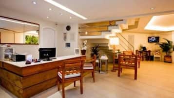 A modern hotel lobby with a reception desk, wooden chairs, a staircase, and a TV mounted on the wall.
